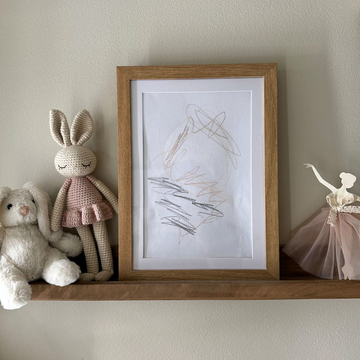 Wooden art frame on a shelf beside soft toys, holding a child's drawing